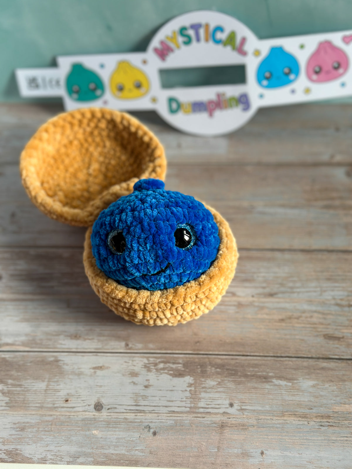 A handmade crocheted amigurumi toy featuring a blue smiling dumpling character sitting inside a yellow half-moon shell, displayed on a rustic wooden surface with a branded sign in the background.