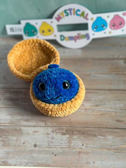 A handmade crocheted amigurumi toy featuring a blue smiling dumpling character sitting inside a yellow half-moon shell, displayed on a rustic wooden surface with a branded sign in the background.