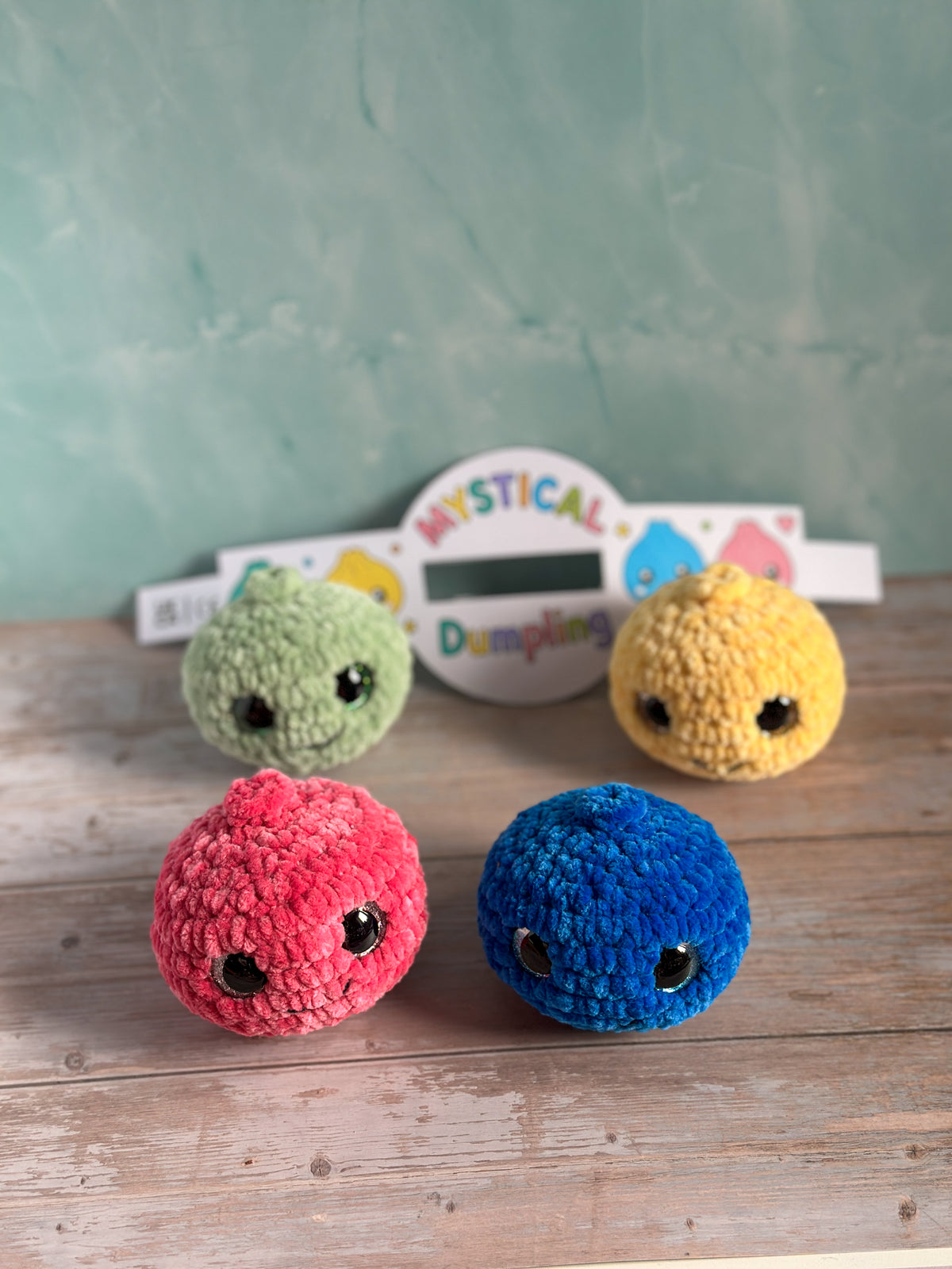 Four small crocheted plush characters in pink, blue, green, and yellow are arranged on a wooden surface in front of a mint green textured wall. A white sign in the background reads 'MYSTICAL Dampifying'.