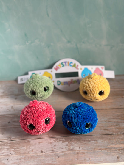 Four small crocheted plush characters in pink, blue, green, and yellow are arranged on a wooden surface in front of a mint green textured wall. A white sign in the background reads 'MYSTICAL Dampifying'.