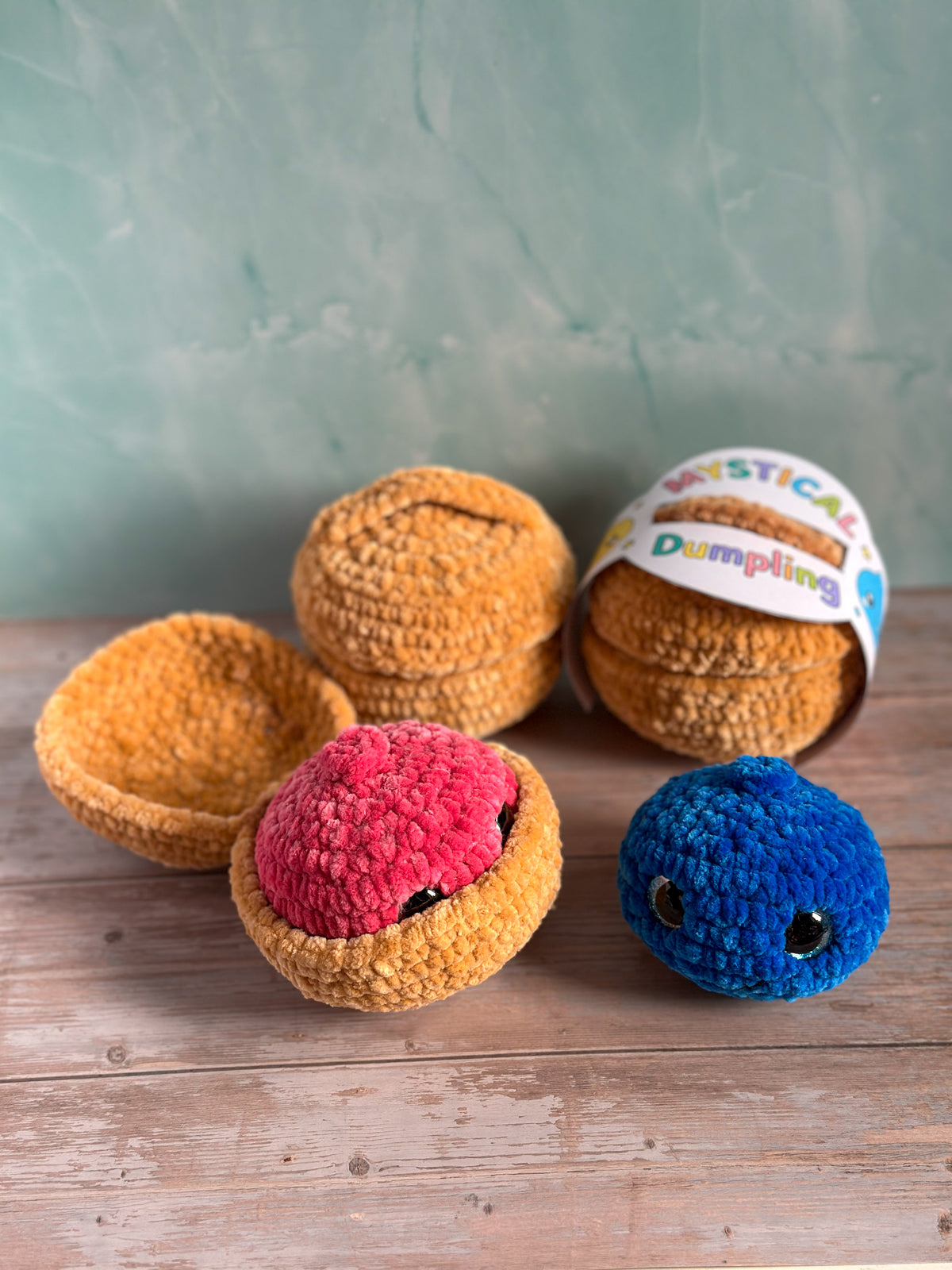 A set of handmade crocheted dumpling toys arranged on a wooden surface against a textured teal wall. The set includes two plain golden-brown dumplings, a pink dumpling with black eyes, a blue dumpling with black eyes, and a packaged brown dumpling with a white paper label that reads 'MYSTICAL Dumpling'.