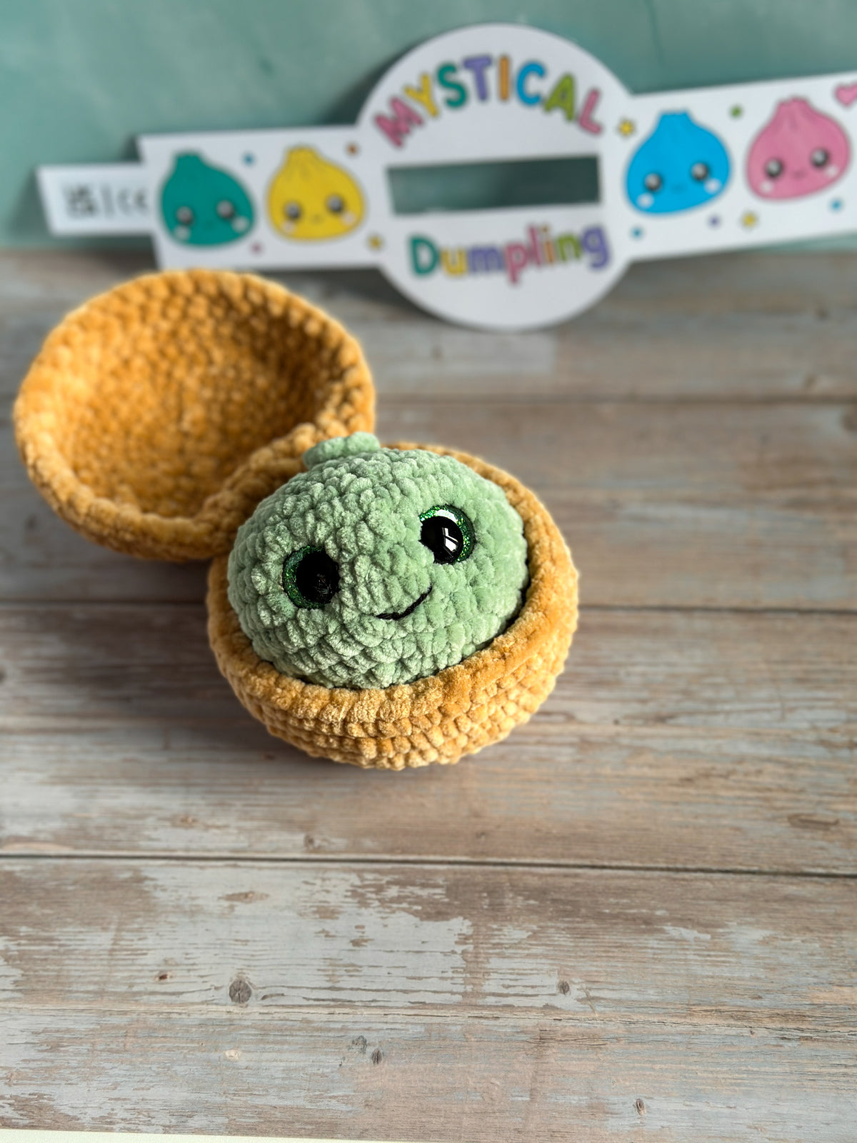 A handmade crochet amigurumi dumpling with a smiling green face sits inside a yellow-orange crocheted bowl on a rustic wooden surface, with a decorative sign reading 'Mystical Dumpling' in the background.