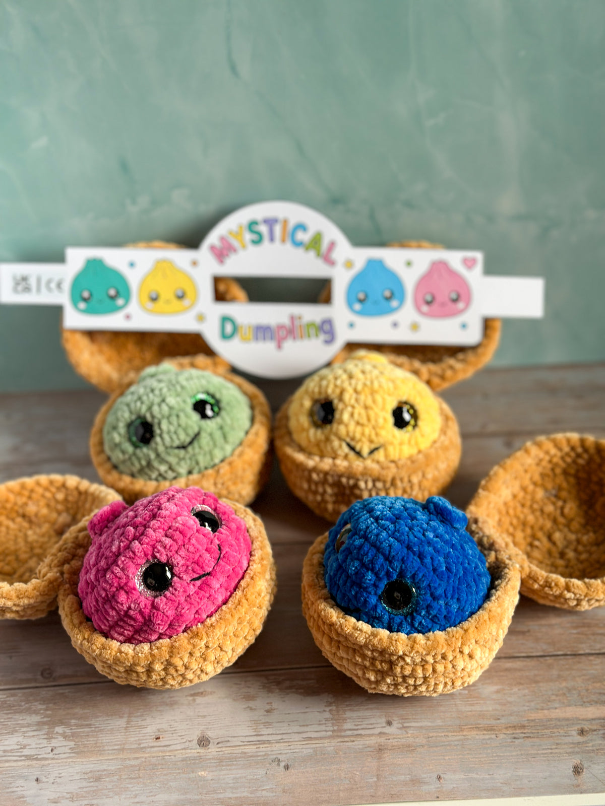 Five crocheted dumpling characters in pink, blue, yellow, and green, each sitting in a small golden-brown basket, arranged on a wooden surface with a branded sign reading 'Mystical Dumpling' in the background.