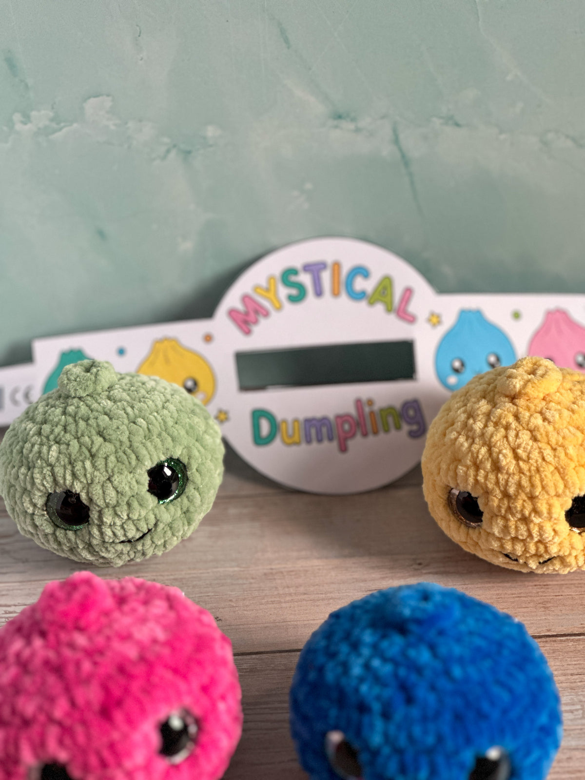 Four knitted dumpling characters in green, yellow, pink, and blue arranged on a wooden surface in front of a white sign that reads 'MYSTICAL Dumpling' against a pale green wall.
