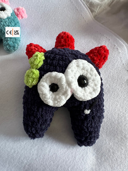 A handmade crocheted monster toy with a dark navy body, large white eyes, red horns, and green spots, resting on a white textured blanket.