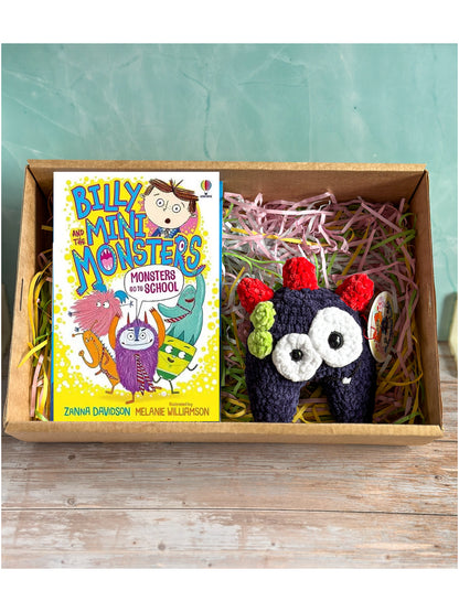 A children's book titled 'Billy and the Mini Monsters: Monsters and School' displayed next to a handmade crocheted monster toy inside a cardboard gift box filled with shredded paper, set against a teal background and rustic wood surface.