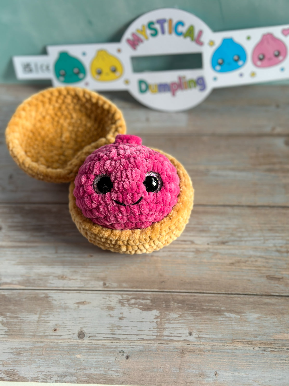 A handmade crochet amigurumi dumpling toy with a pink filling and yellow shell, displayed on a rustic wooden surface with a branded sign in the background.