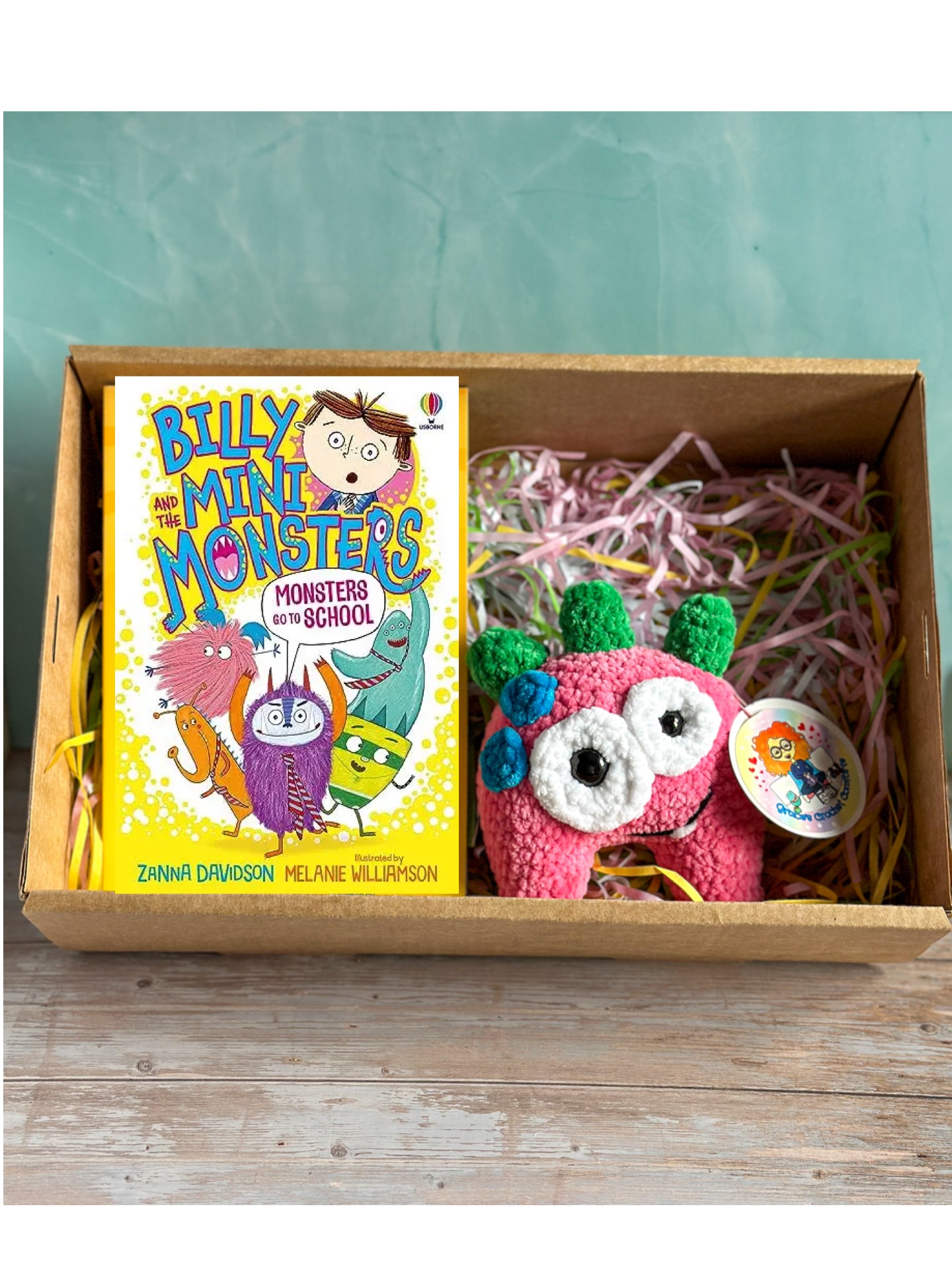 A children's gift box set containing the book 'Billy and the Monsters: Monsters with School' by Zainab Dawison and Melanie Millippon, placed next to a pink handmade monster plush toy with green ears and a round sticker, all arranged in a cardboard box with shredded paper filler.