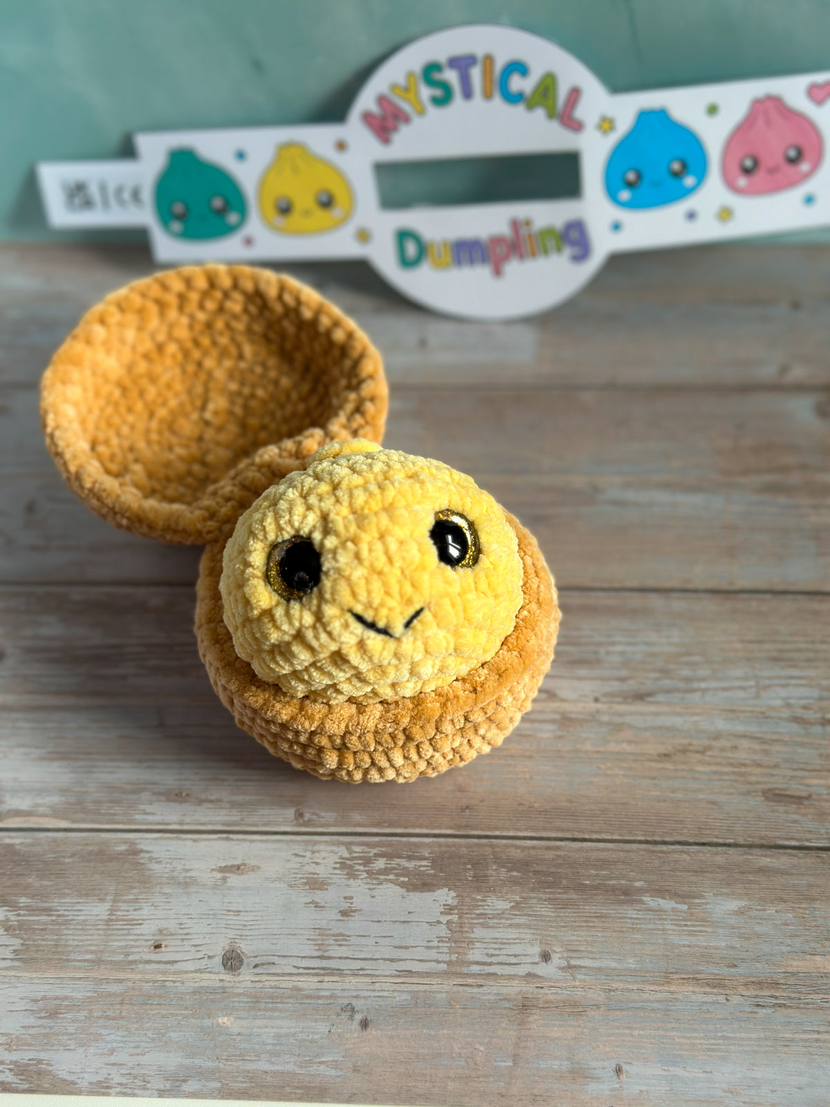 A handmade crocheted yellow smiling dumpling toy with a matching hollow dumpling cup, displayed on a rustic wooden surface with a branded sign in the background.
