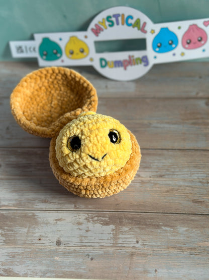 A handmade crocheted yellow smiling dumpling toy with a matching hollow dumpling cup, displayed on a rustic wooden surface with a branded sign in the background.